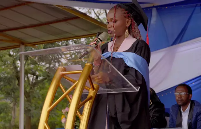 A student in graduation attire speaking at a podium during a ceremony