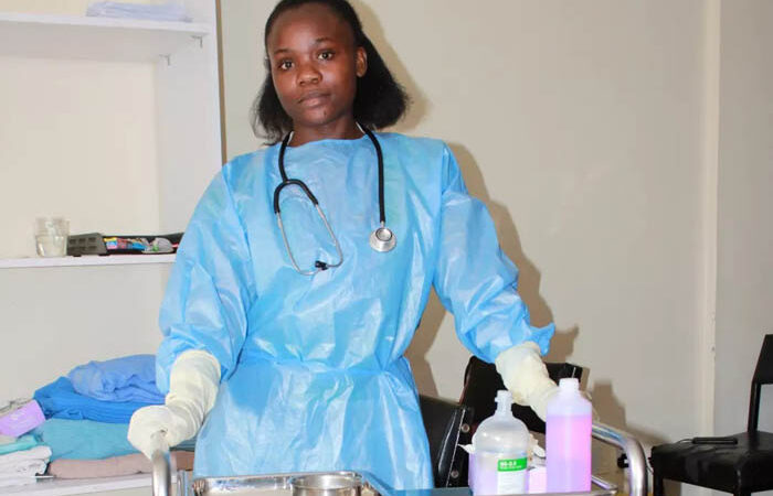 A female medical student in full personal protective equipment, including a blue gown and gloves, with a stethoscope around her neck